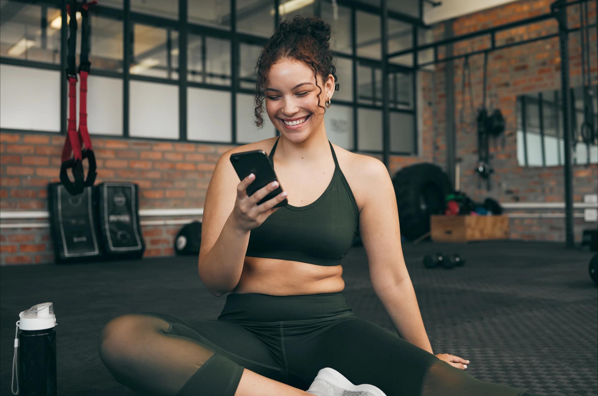 Mujer sentada sonriendo y observando su celular. Enfrente hay una botella de agua, detrás hay un espejo y distintos artefactos para ejercitarse.