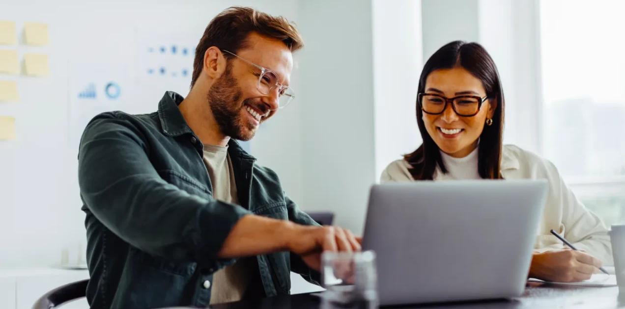 A imagem tem duas pessoas, um homem e uma mulher, eles estão olhando para o notebook, enquanto trabalham.