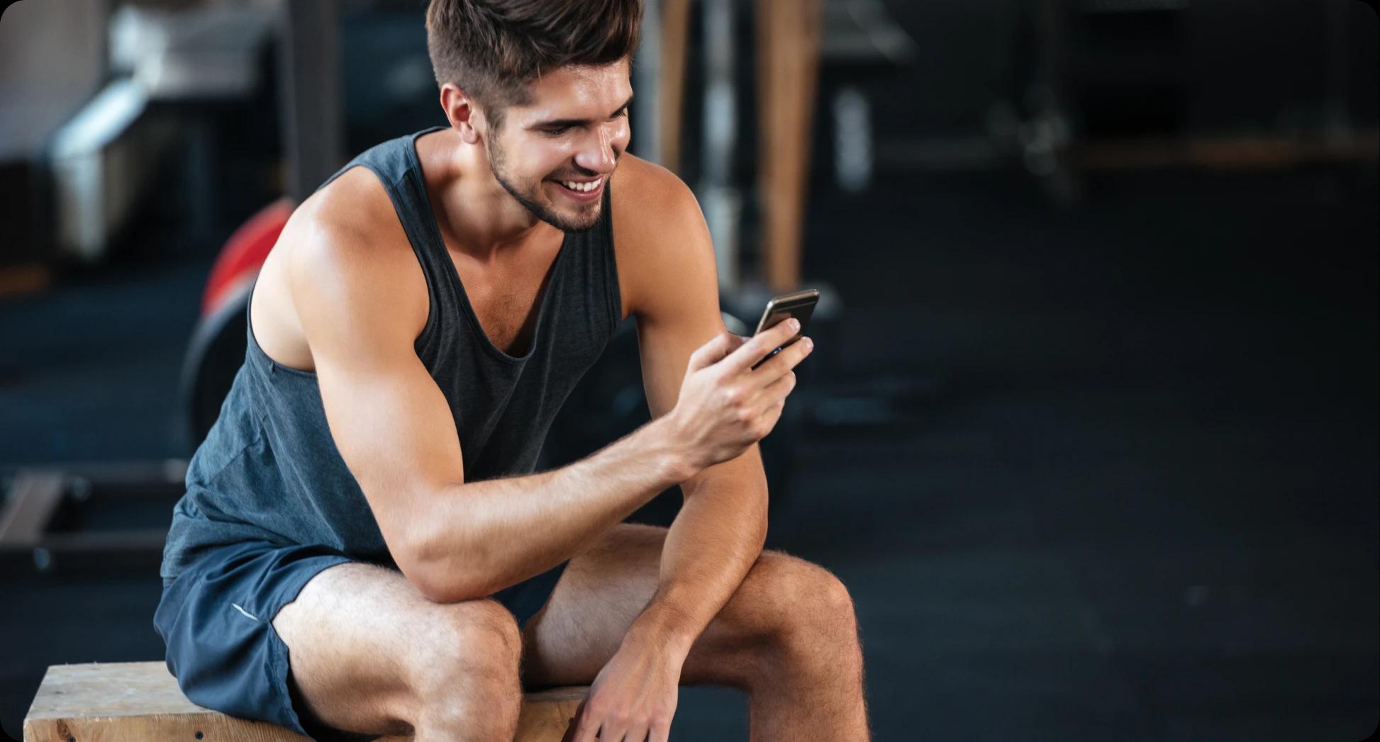 Hombre sentado con ropa deportiva y celular en mano accediendo a su app TotalPass para entrenar. Al fondo se ven aparatos fitness muy desenfocados.