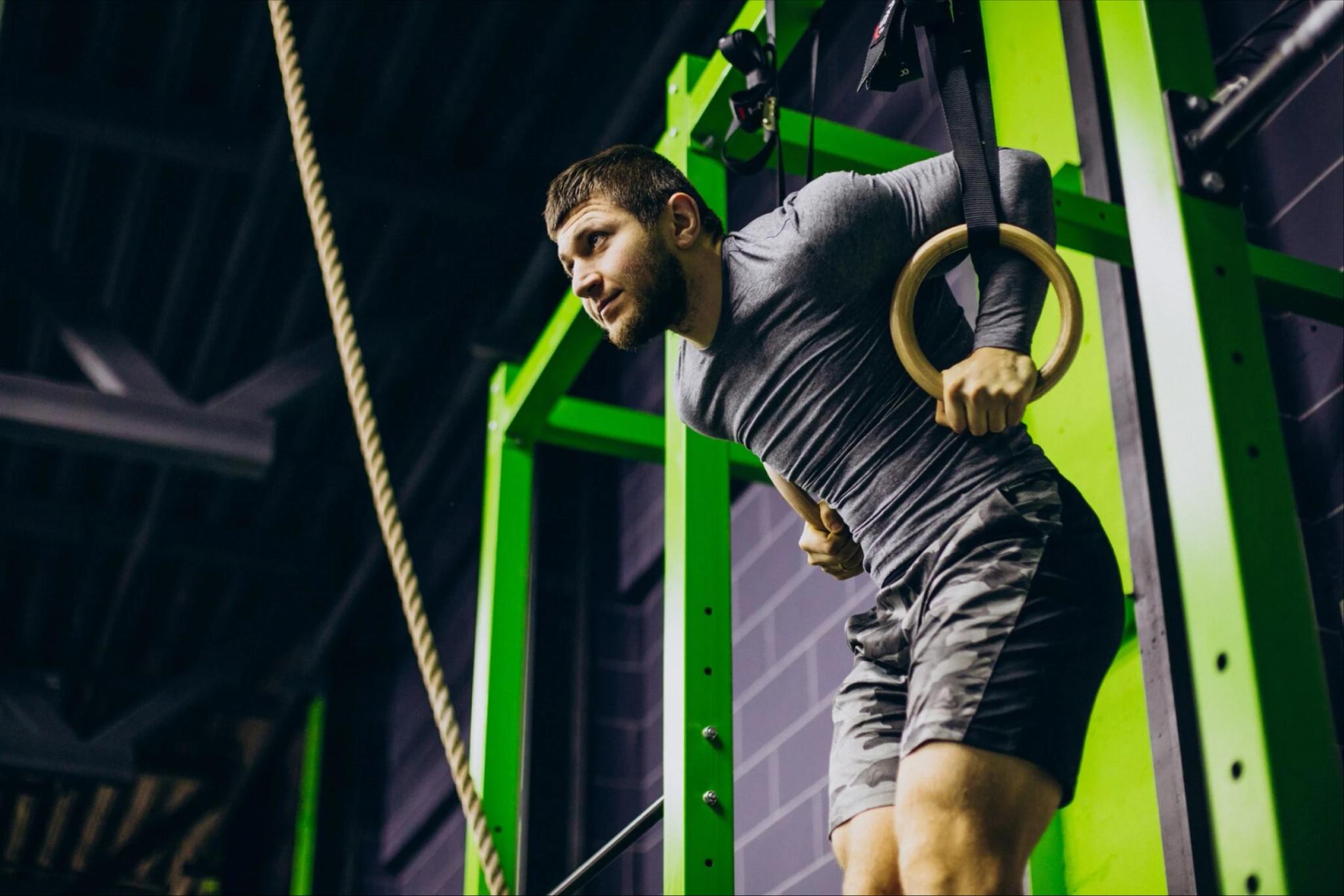 Hombre colgado sosteniendo dos anillos de gimnasia. Su espalda se encuentra levemente inclinada hacia adelante y se observa una cuerda frente a él.