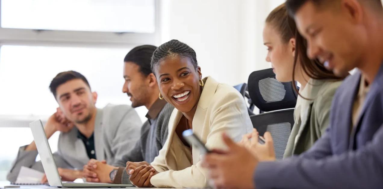 A imagem tem vários colaboradores trabalhando dentro da empresa, tanto homens quantos mulheres. Na mesa, tem notebooks e celulares.