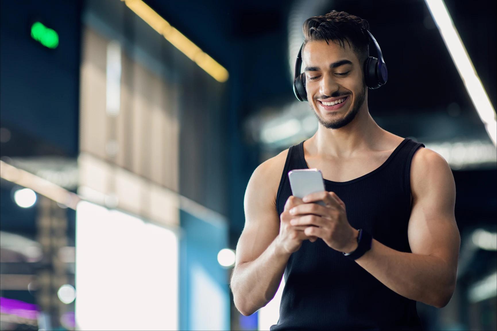 Hombre con audífonos y reloj, sosteniendo su celular con las dos manos mientras lo observa y sonrie. Al fondo se ven lámparas encendidas y cristales.