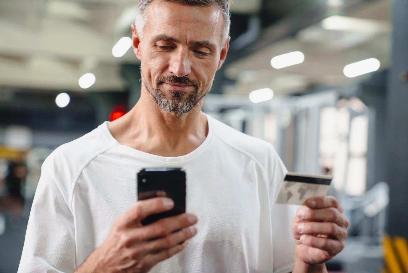 A imagem tem um homem mais velho mexendo no celular. Ele está na academia. 