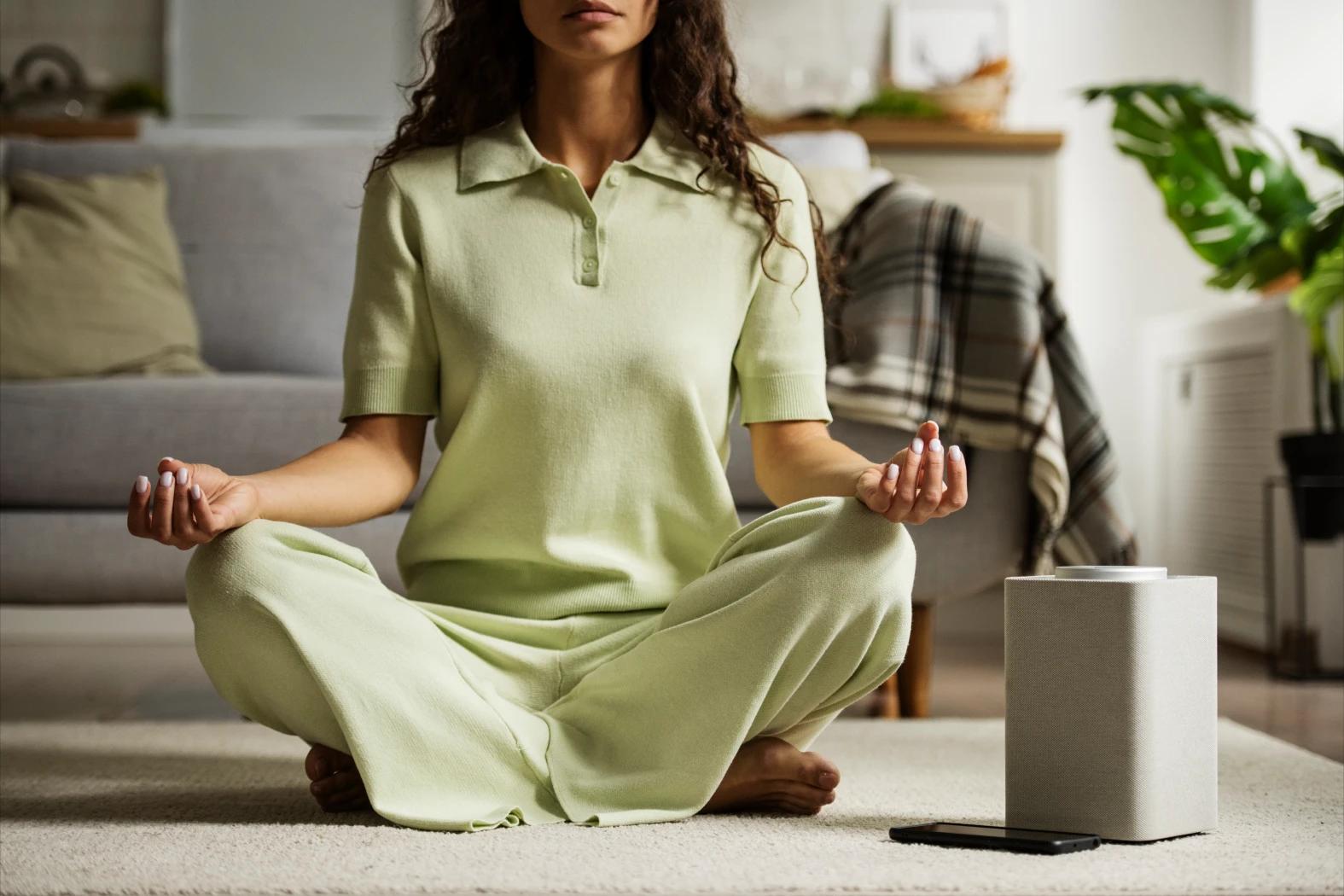 Mujer sentada con las piernas cruzadas y sus manos sobre ellas en posición de meditación. Su celular está en frente y al fondo se observa un sillón.