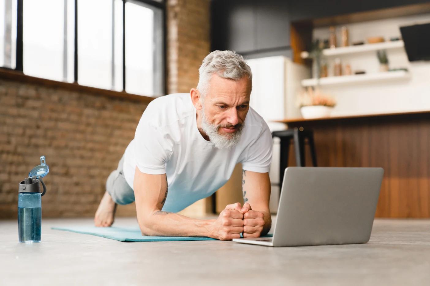 A imagem tem um homem mais velho treinando. Ele está fazendo o exercício prancha, enquanto olha para o notebook.