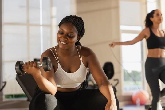 A imagem tem uma mulher com roupa de ginástica. Ela está segurando o halter com o braço direito e exercitando-se.