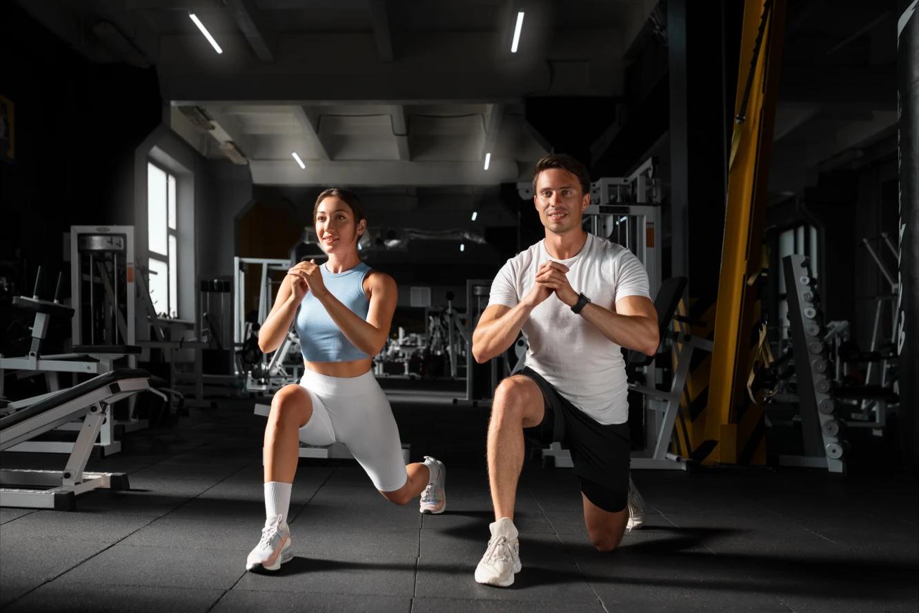 Hombre y mujer haciendo desplantes en un gimnasio. Ambos están sonriendo y detrás de ellos se observan aparatos para hacer ejercicio.