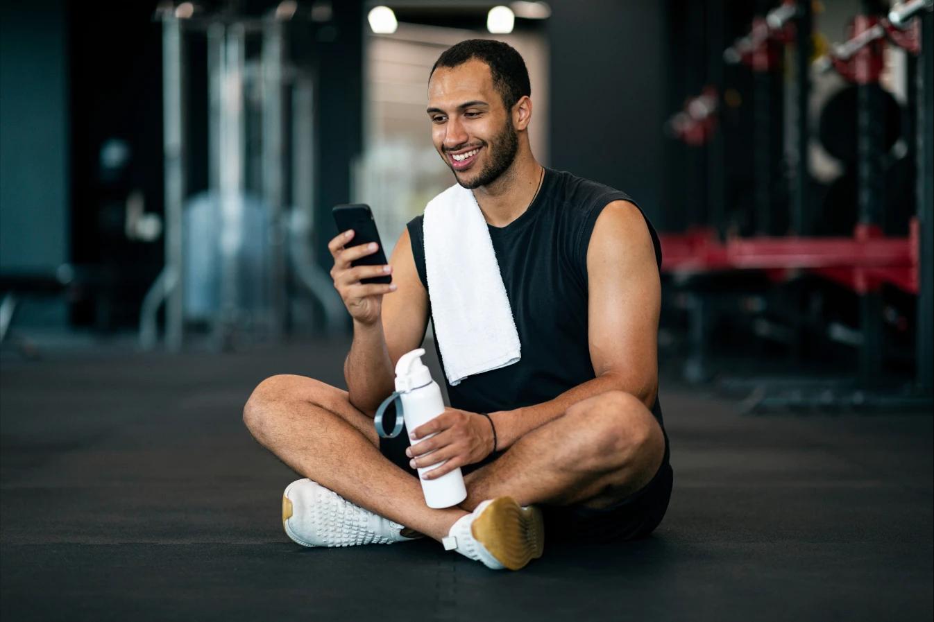 Hombre sentado en un gym con los pies cruzados, una toalla, botella de agua y su celular en mano. Al fondo se ven aparatos de ejercicio desenfocados