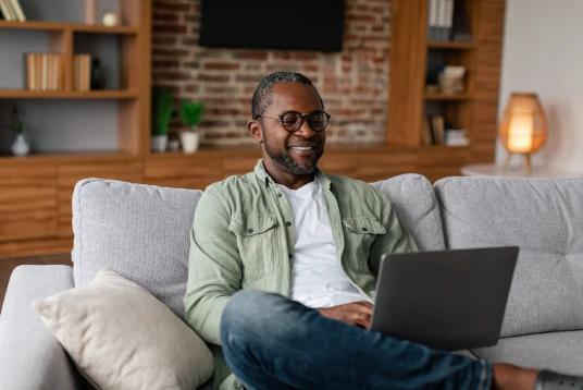 A imagem tem um homem mais velho, mexendo no notebook e sentado no sofá da sala.