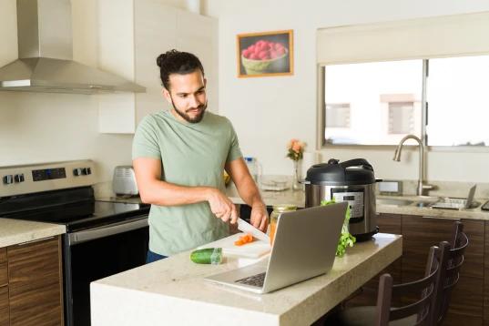 A imagem tem um homem mais jovem cozinhando. Ele está vendo a receita pelo notebook, enquanto corta legumes.