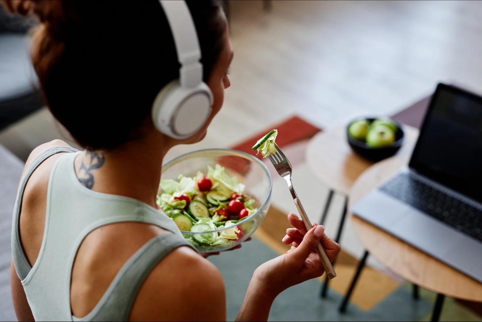 Mujer con audífonos comiendo una ensalada verde frente a una computadora para mantener una alimentación equilibrada y nutritiva.