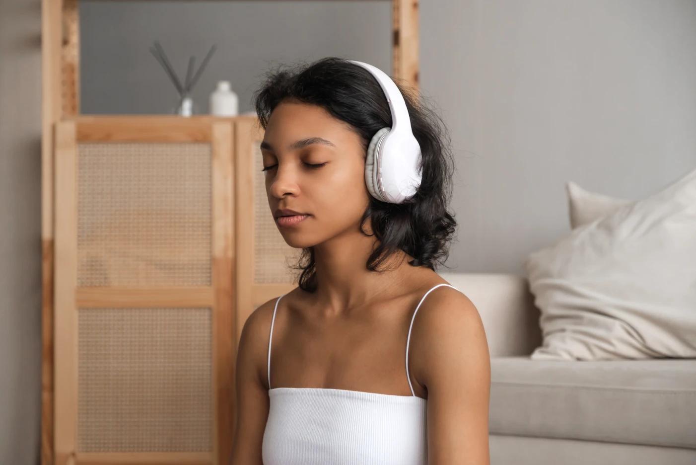 A imagem tem uma menina com headphone. Ela está no seu quarto, concentrada e de olhos fechados. 