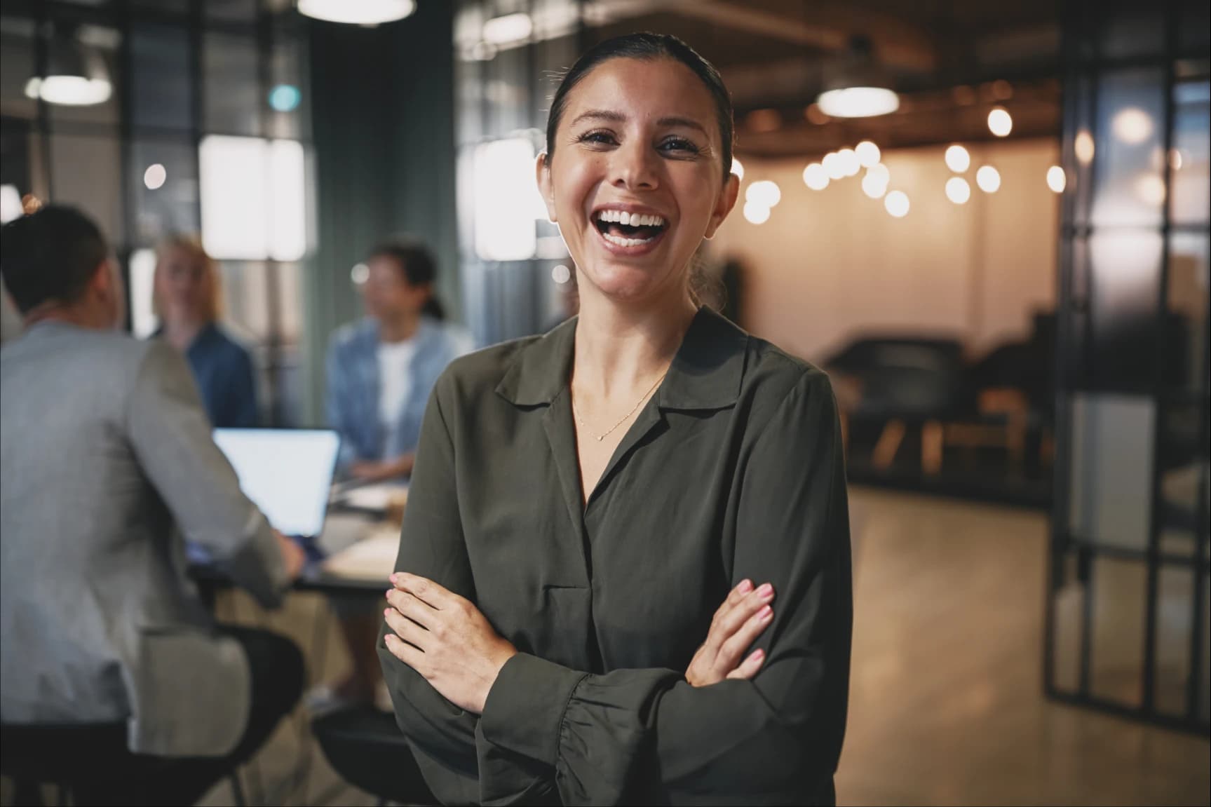 Mujer sonriente en una oficina Mujer riendo con los brazos cruzados dentro de una oficina. Al fondo se ven otros colaboradores sentados en un escritorio con una computadora.