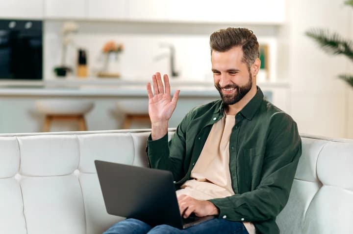 Starbem A imagem tem um homem de meia-idade sentado no sofá, sorrindo e acenando para a câmera de um notebook.