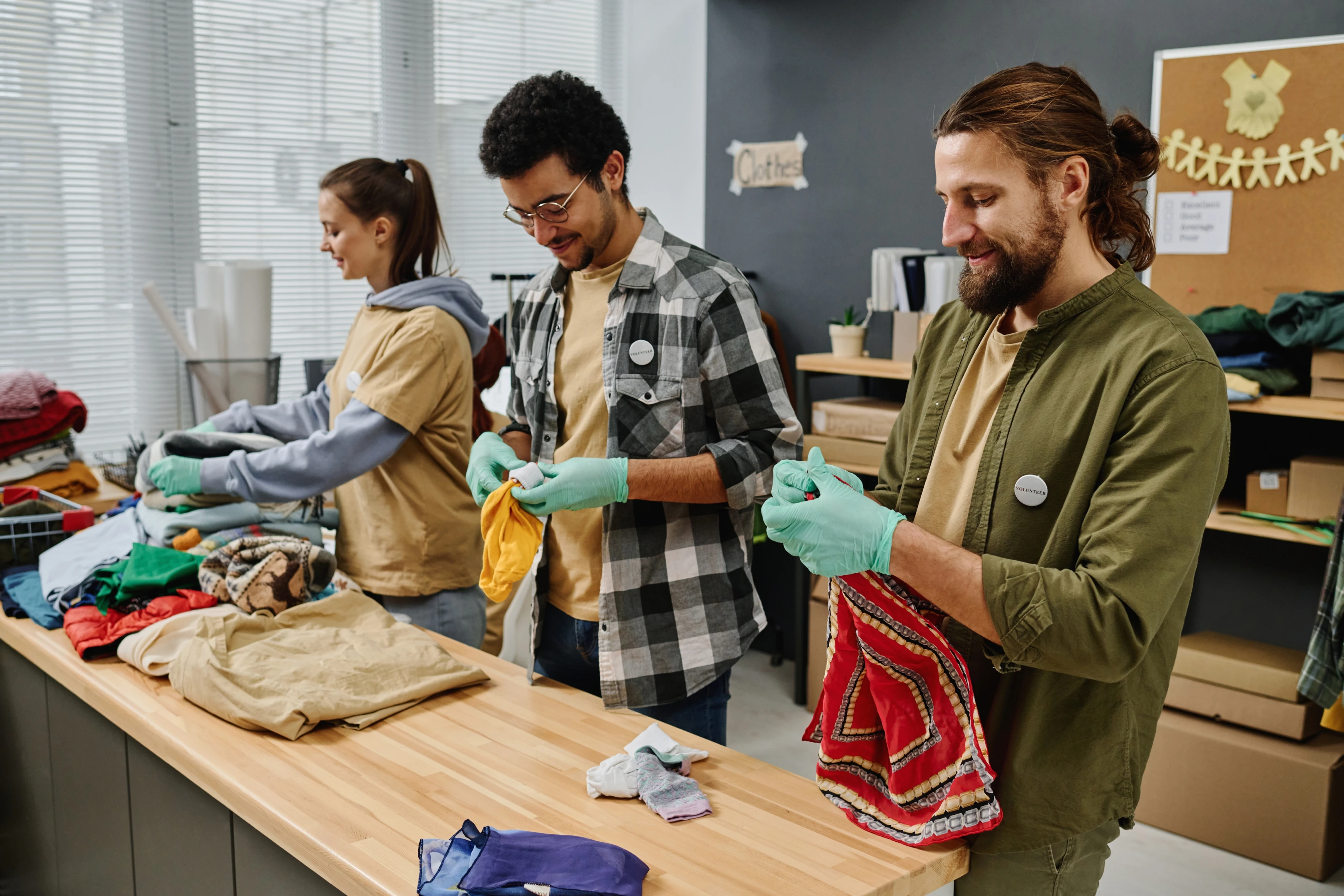 A imagem tem três pessoas, dois homens e uma mulher. Ambos estão dobrando e organizando as roupas arrecadadas.