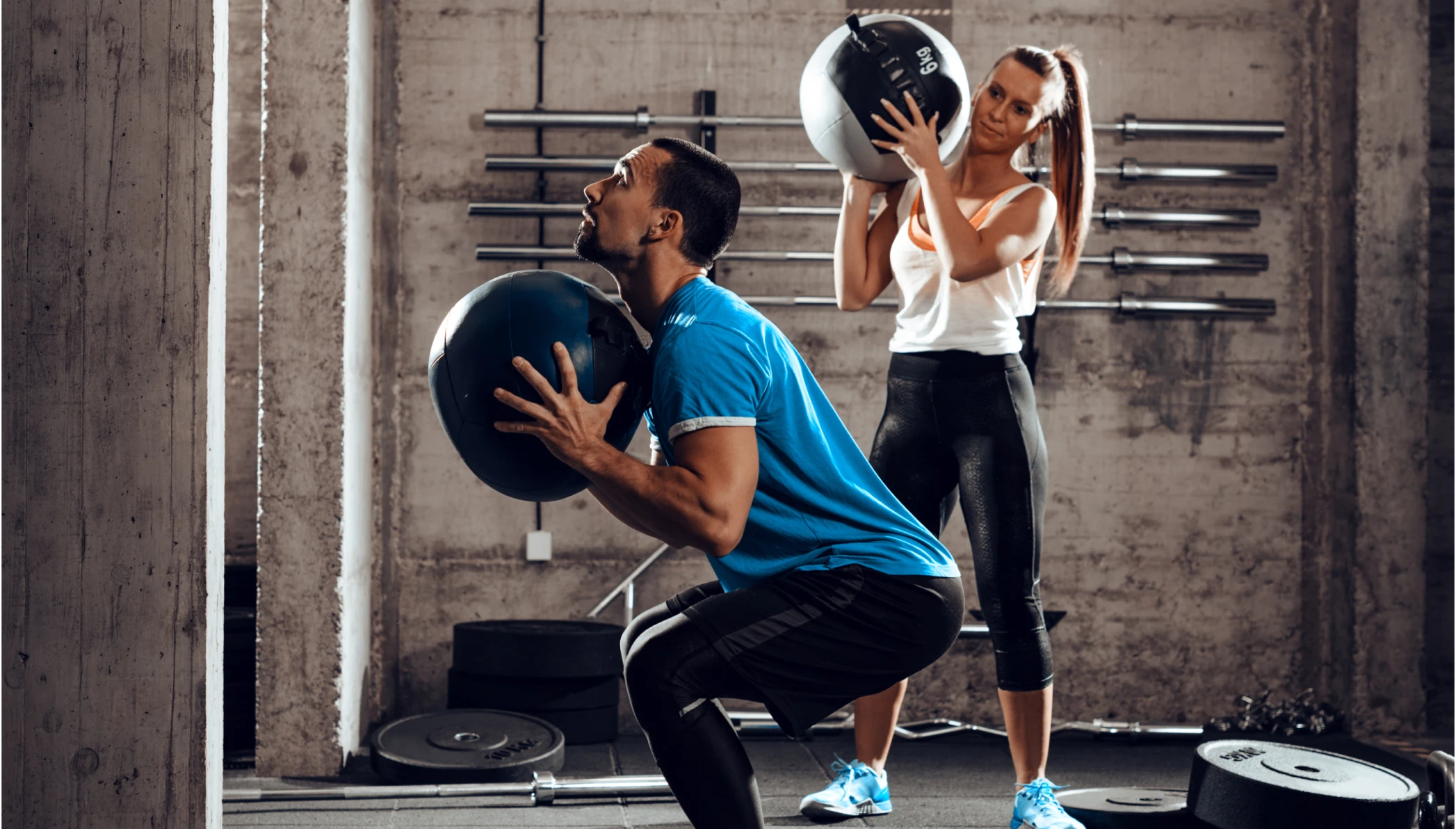 Hombre y mujer practicando pilates con pelota en mano Hombre con piernas flexionadas y una mujer parada detrás de él. Ambos sostienen una pelota grande y en el suelo se observan pesas para ejercitarse.
