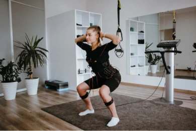 Na foto, há uma mulher enquadrada ao centro, que está agachada, pisando em um tapete, realizando um dos exercícios do treino de eletroestimulação. Nela, há alguns equipamentos, além de vestir uma roupa própria para a prática do exercício. #ParaTodosVerem