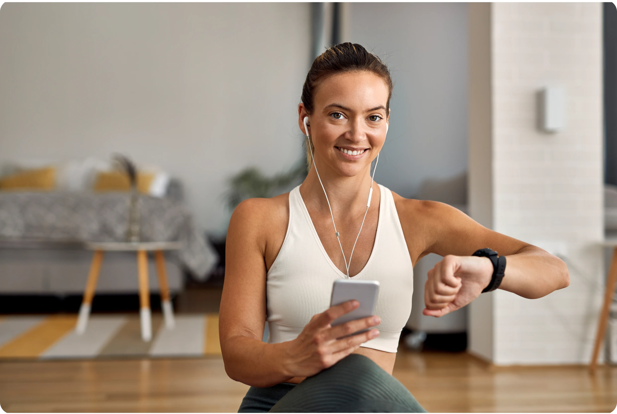 Mujer con ropa deportiva, audífonos y reloj mira al frente y sonríe mientras sostiene su celular. Detrás hay un sofá, un tapete y una mesa de centro.