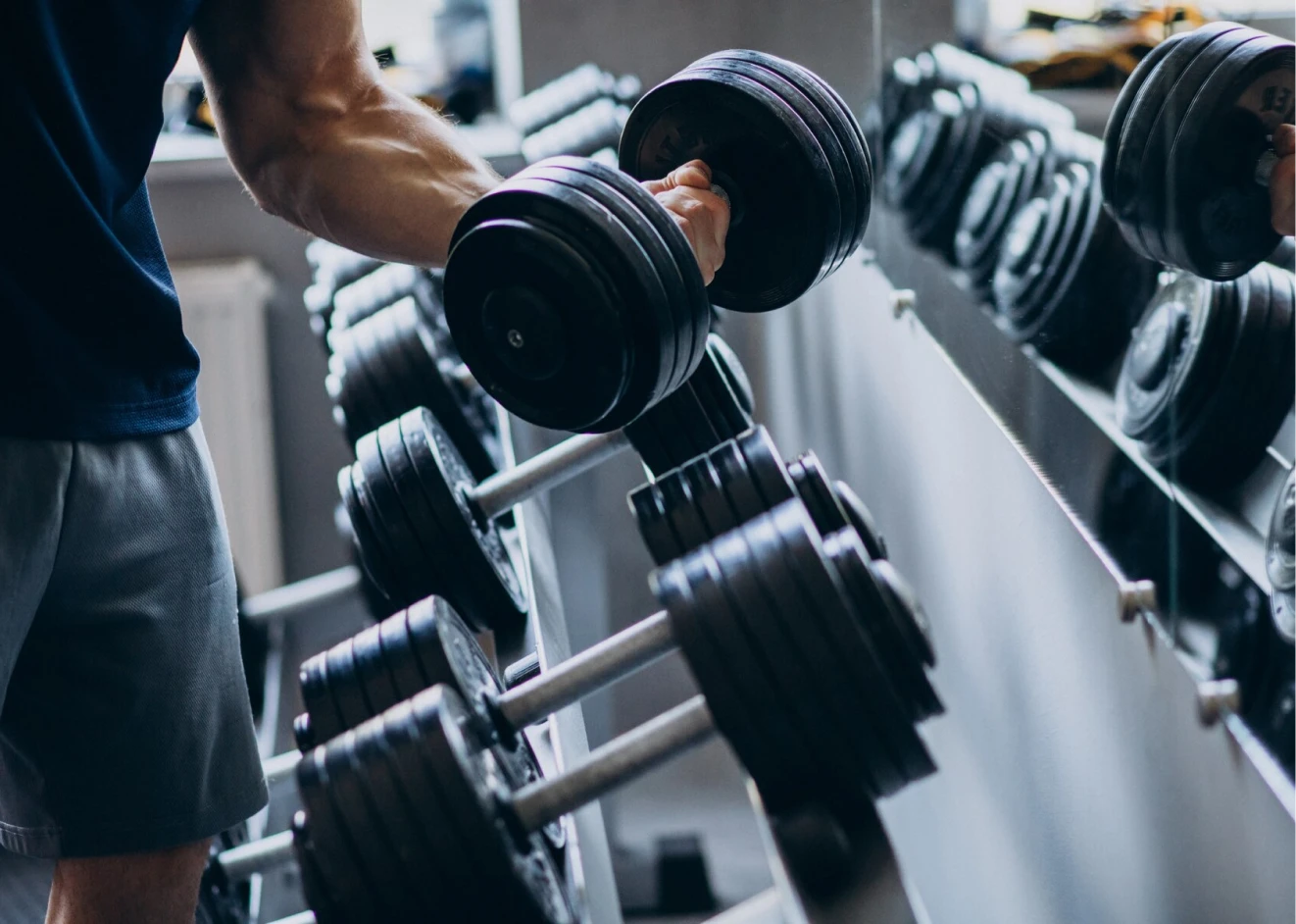 La imagen muestra únicamente la parte del abdomen y el muslo de un hombre que con su brazo está sosteniendo una pesa frente al espejo de un gimnasio.