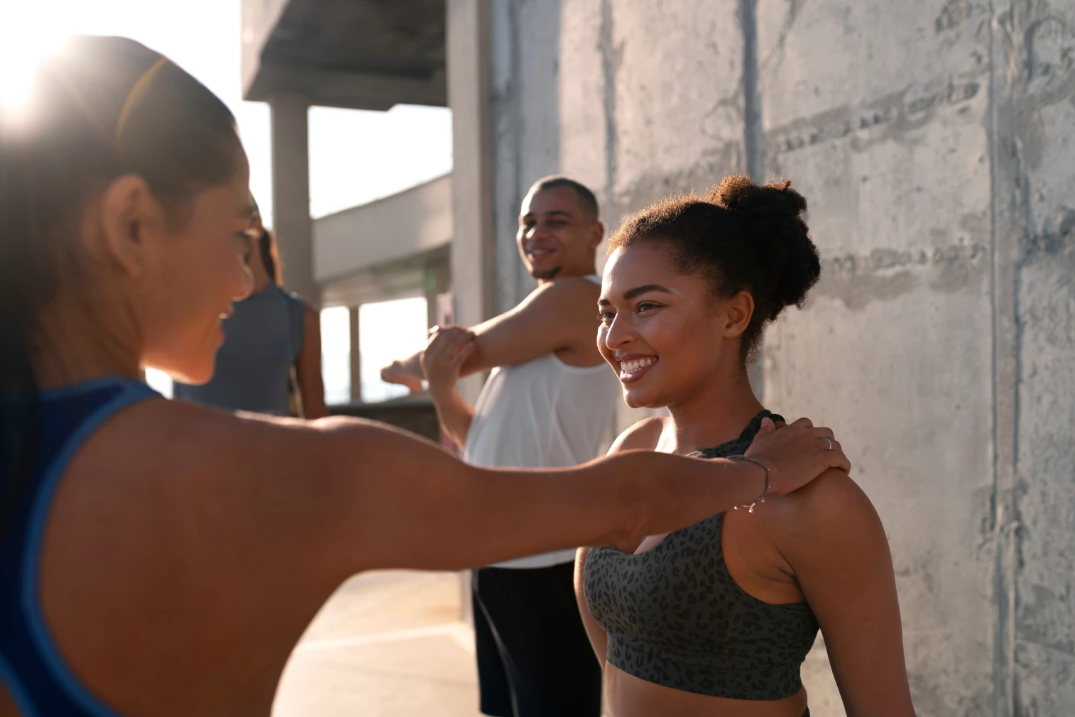 Dos mujeres se observan de frente, mientras una toca los hombros de la otra con sus brazos estirados. Un hombre las observa sonriendo mientras estira.