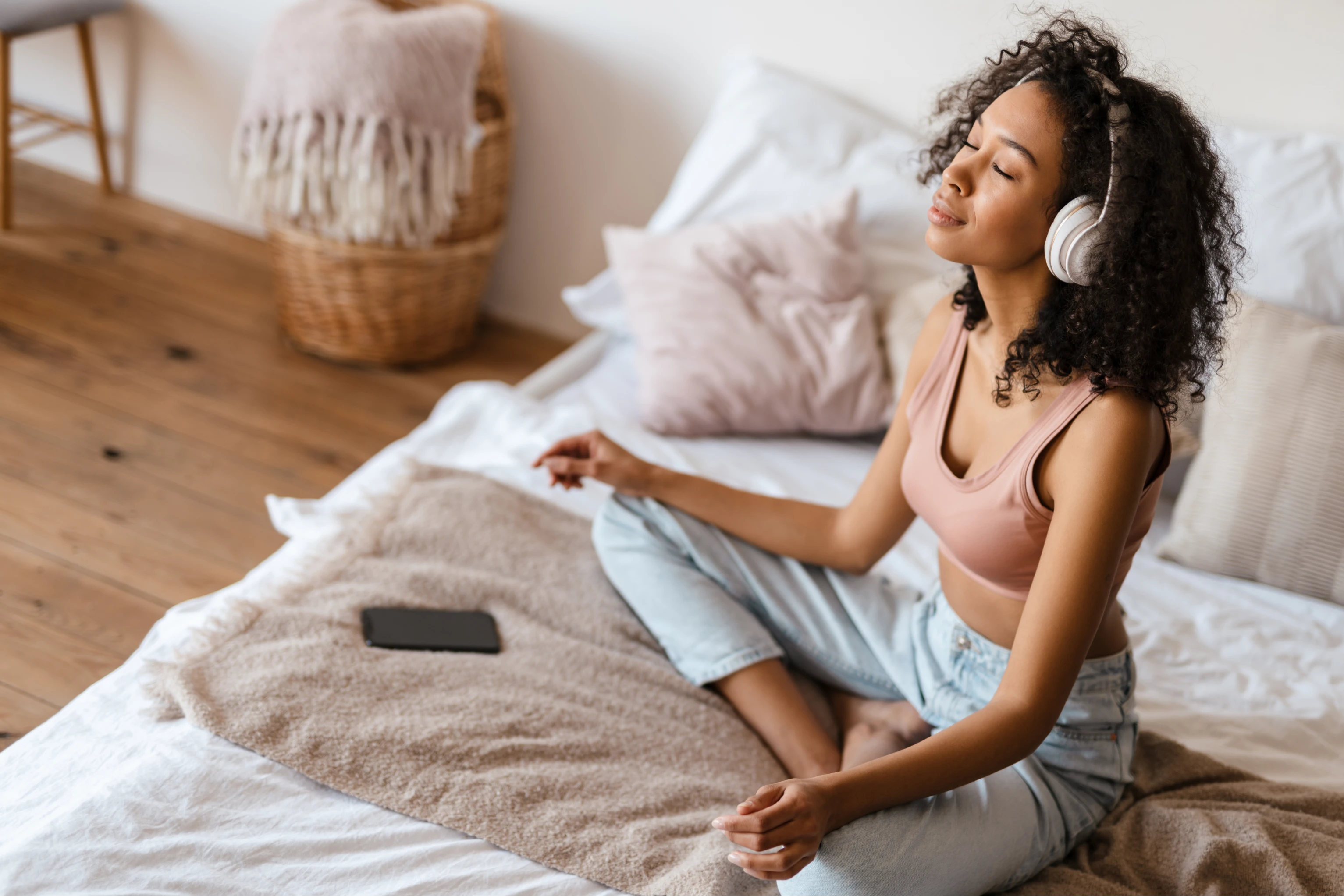 A imagem tem uma mulher meditando. Ela está com um headphone e está sentada com as pernas cruzadas em cima da cama.