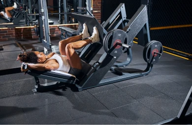 A foto se passa em uma academia. Nela, há uma mulher realizando um exercício de elevação para os membros inferiores. #ParaTodosVerem