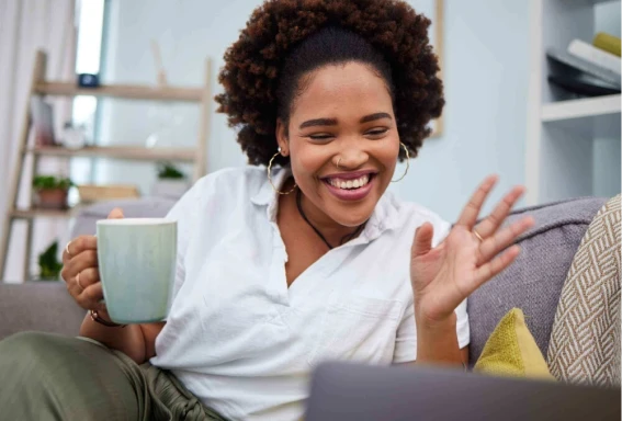 Uma mulher em uma sala, sentada no sofá, sorrindo e segurando uma xícara, utilizando seu notebook à frente, como se estivesse em uma chamada de vídeo. #ParaTodosVerem