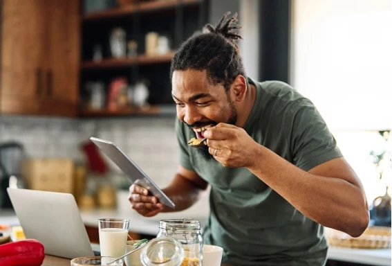 Um homem na cozinha de casa, fazendo uma refeição, enquanto segura um tablet. Em frente, há uma mesa com alguns alimentos, como nozes, leite e frutas. #ParaTodosVerem