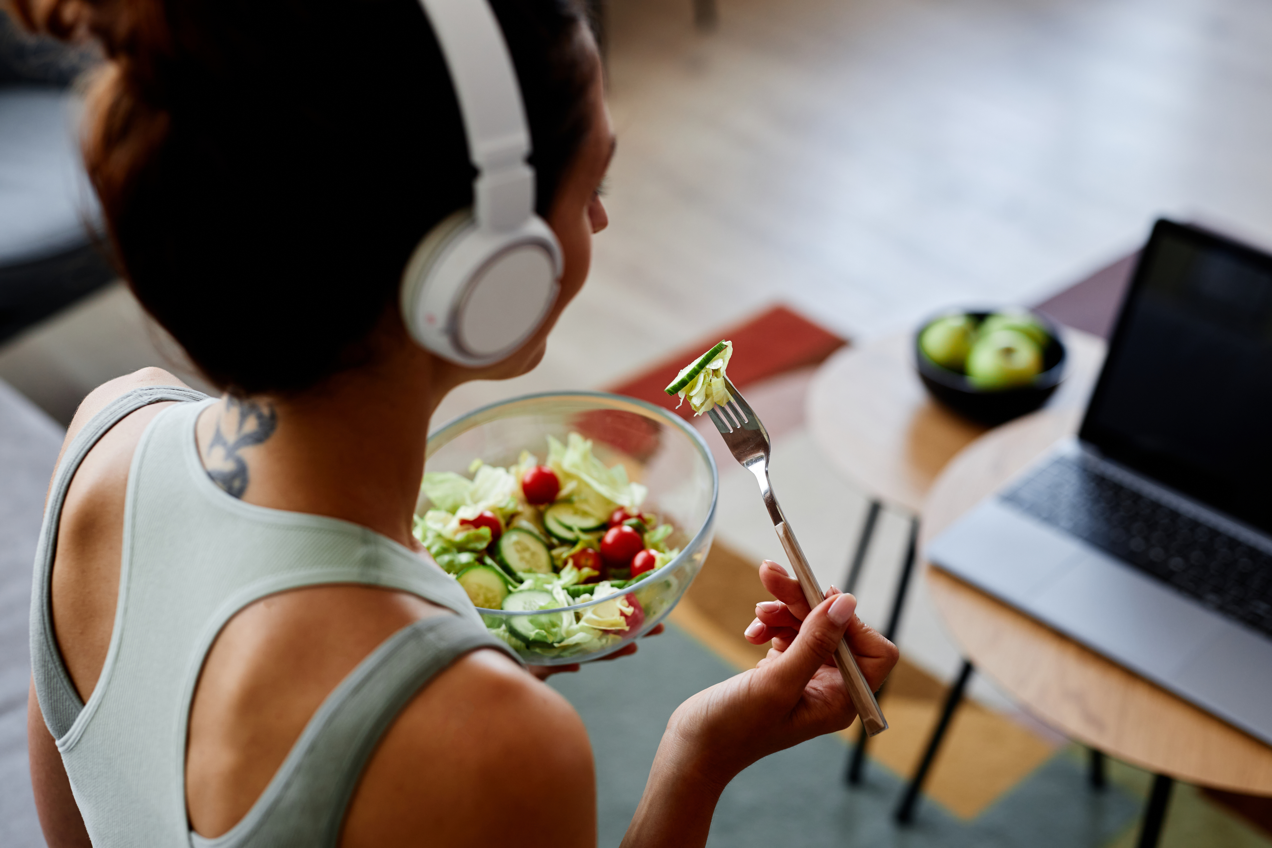 Mujer con top deportivo, audífonos y frente a una computadora, comiendo una ensalada nutritiva para cuidar de su bienestar integral.