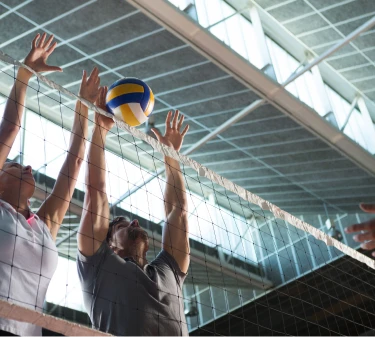 A imagem mostra três pessoas praticando vôlei. A foto foi feita em um ginásio, e duas das três pessoas estão fazendo o bloqueio da bola, esticando os braços acima da rede. #ParaTodosVerem