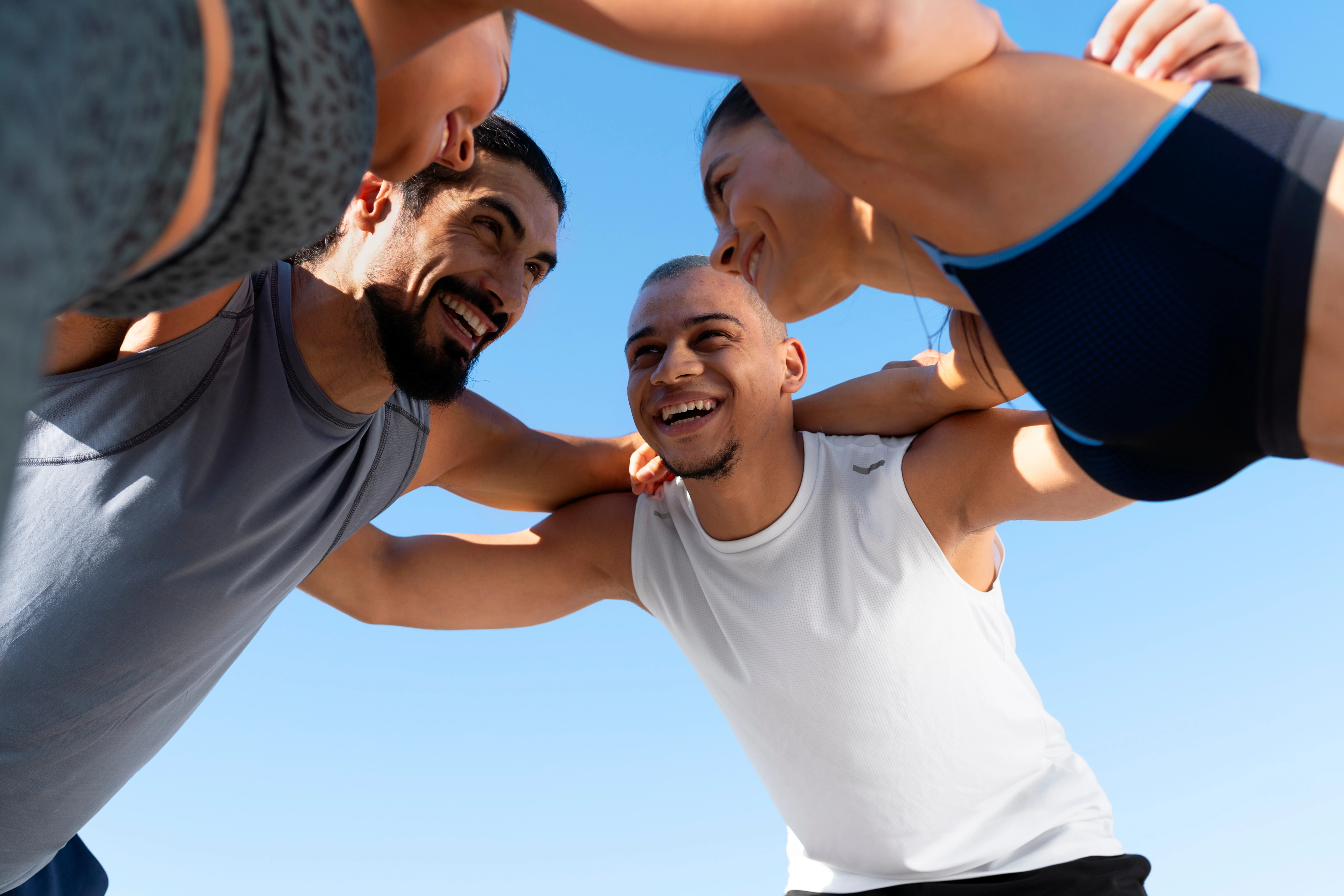 Dos mujeres y dos hombres abrazados con ropa deportiva, están ligeramente inclinados y formando un círculo con sus brazos. De fondo se ve el cielo.